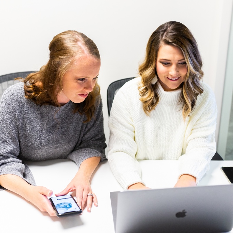 Two people working through an app together, one holding a phone, the other at a laptop