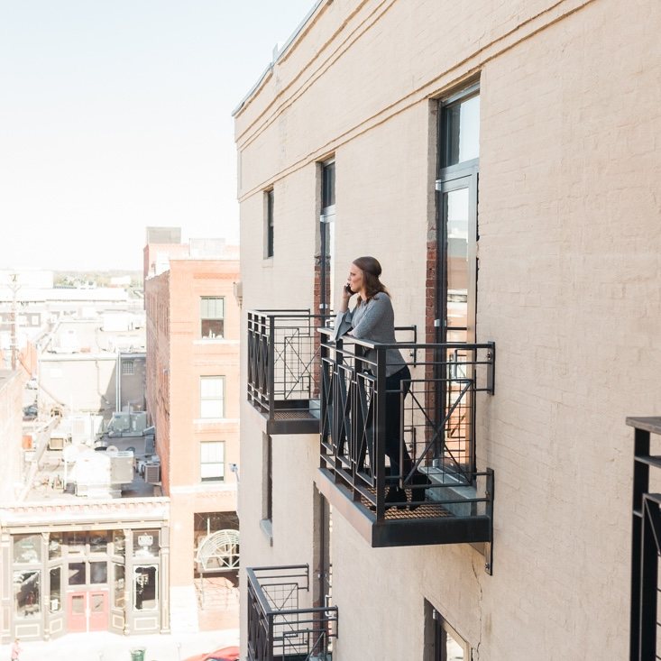 Becca on a fire-escape balcony several storeys above a downtown street, talking on the phone