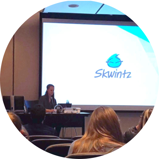 Becca presenting to a seated audience, a projection screen behind her showing the Skwintz logo
